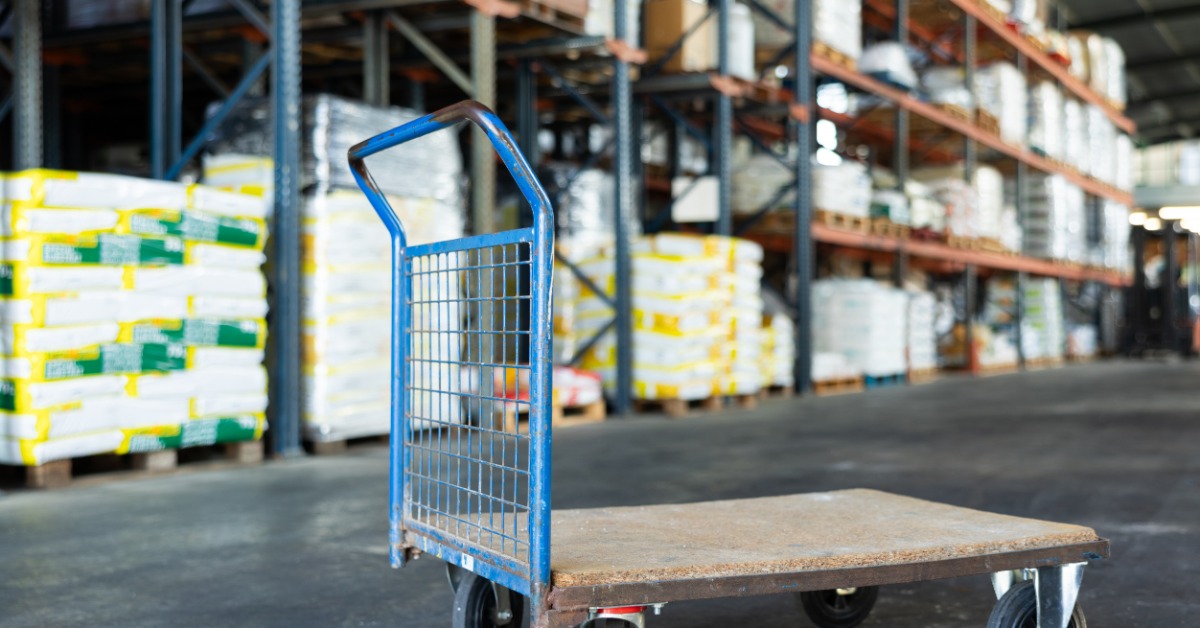 A warehouse cart with a wooden bottom and a large, blue handle. Behind it is a row of warehouse racking.