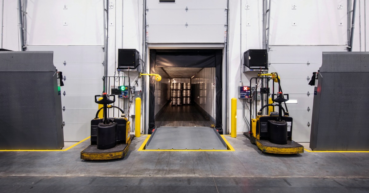 The inside of a large, commercial warehouse facility with an open loading dock door. There are forklifts next to the door.