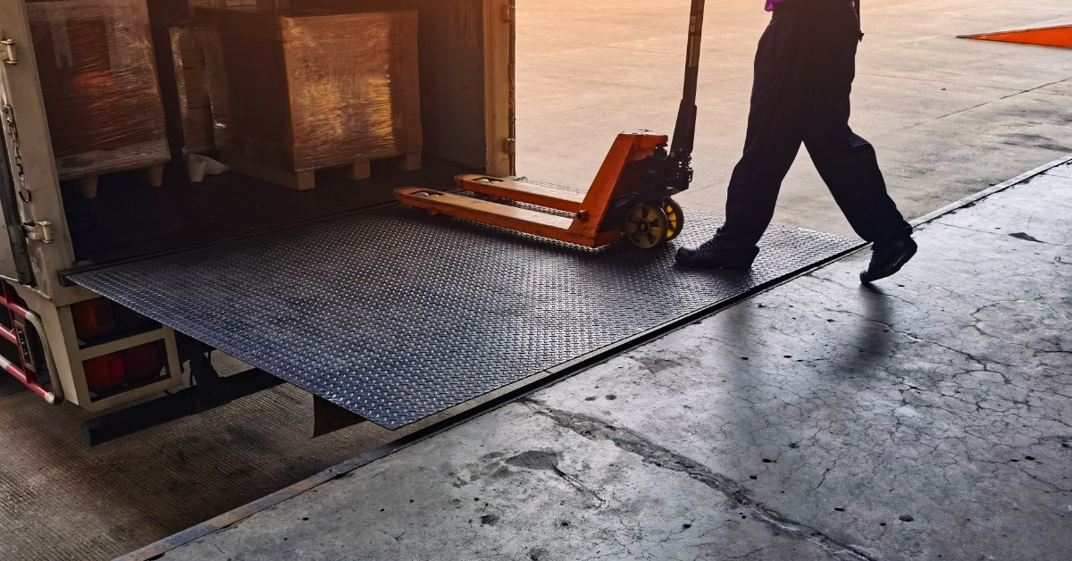 A close-up of a warehouse employee using a pallet jack to unload a truck at the loading dock. The jack has two arms.