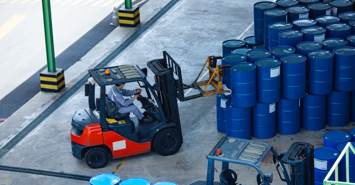 An aerial view shows a person using a red and black forklift to grab a blue drum from a stack of drums.
