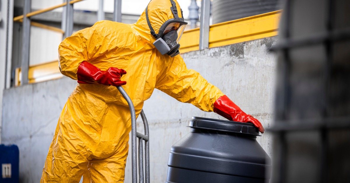 A person dressed in a yellow safety suit, gas mask, and red gloves is moving a black drum with a moving dolly.